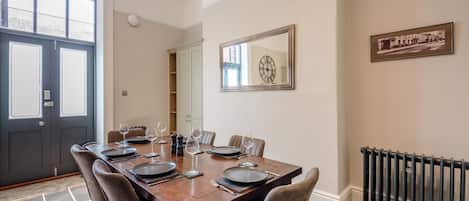 A dining room with a large table set for six, a black door, a clock on the wall, and a radiator.