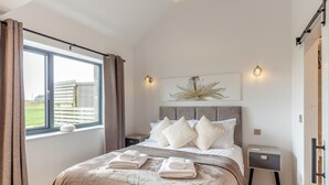 Un dormitorio con cama, almohadas, mesita de noche, cortinas en la ventana y una lámpara de pared.