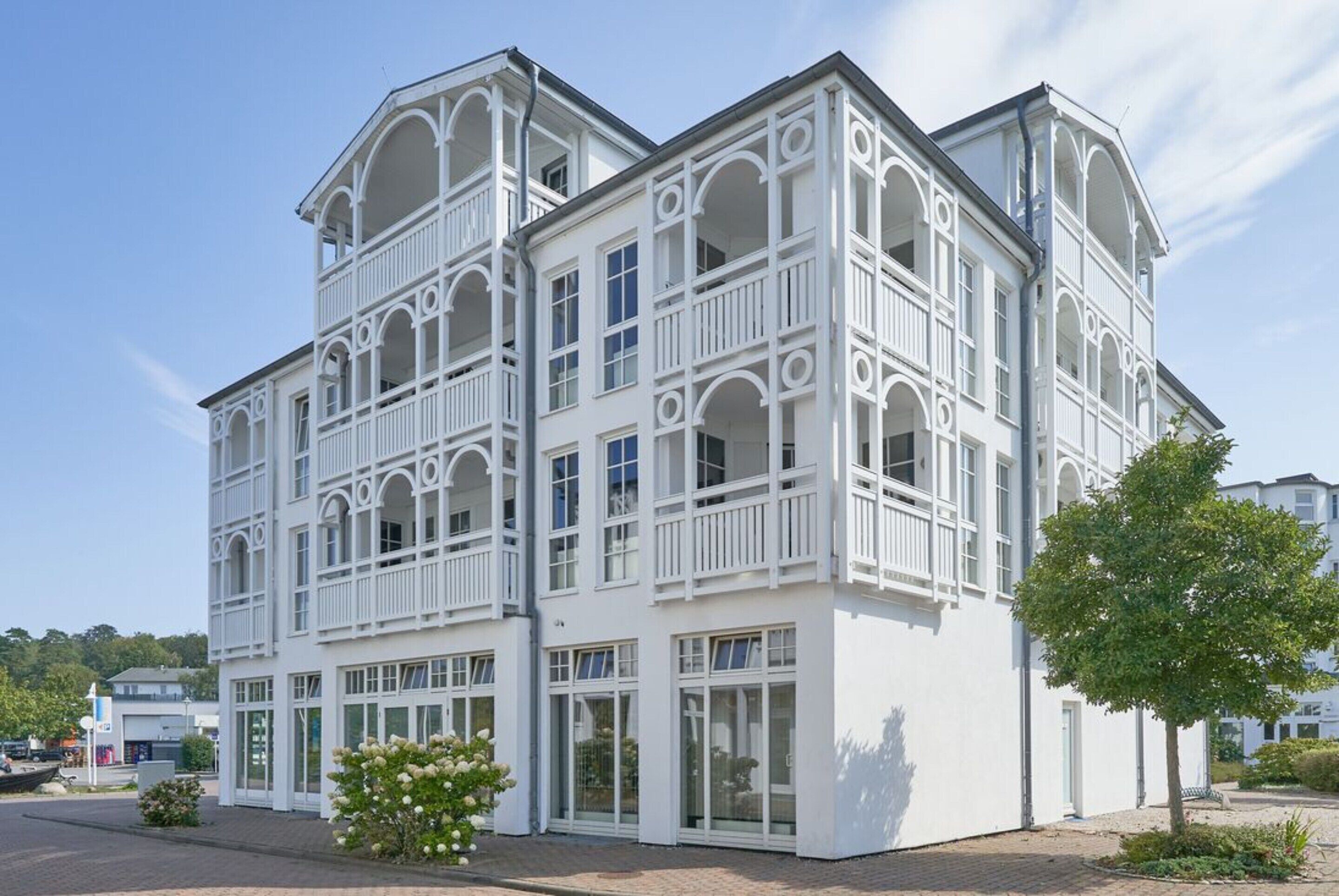 A white building with multiple balconies and large windows, surrounded by a paved area and a tree.