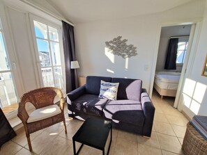 A living room with a sofa, a chair, a small table, a lamp, and a bed. - Ferienwohnung mit Balkon in Sonniger Sud-west-lage in Sellin (Sellin)