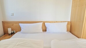 A hotel room with a wooden headboard, two white beds, a bedside table with a lamp, and a power outlet. - Ferienwohnung mit Balkon in Sonniger Sud-west-lage in Sellin (Sellin)