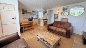 A cozy living room with a brown leather sofa, a wooden coffee table, and a dining table with benches. - Coastal Cottage Retreat Steps from the Beach - Pass the Keys (Kirkcudbright)