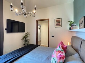 A bedroom with a bed, a TV mounted on the wall, a chandelier, a mirror, a door leading to another room, and a shelf with books and a plant. - Da Vittoria Lovely apt near the Arena (Verona)