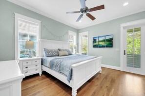 Un dormitorio con cama blanca, suelo de madera, ventilador de techo y vistas a zonas verdes.