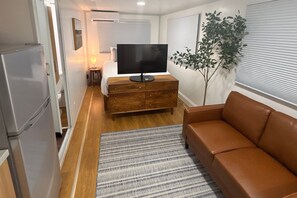 A compact living space with a sofa, a television on a wooden dresser, a small table with a lamp, and a potted plant.