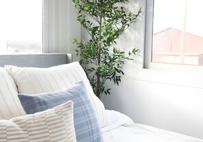 A bed with white bedding and blue and white pillows, a plant beside the window, and a view of a building outside. - Private Duplex Studio Suite with Queen Bed, Kitchenette and Private Bathroom. (Pecos)