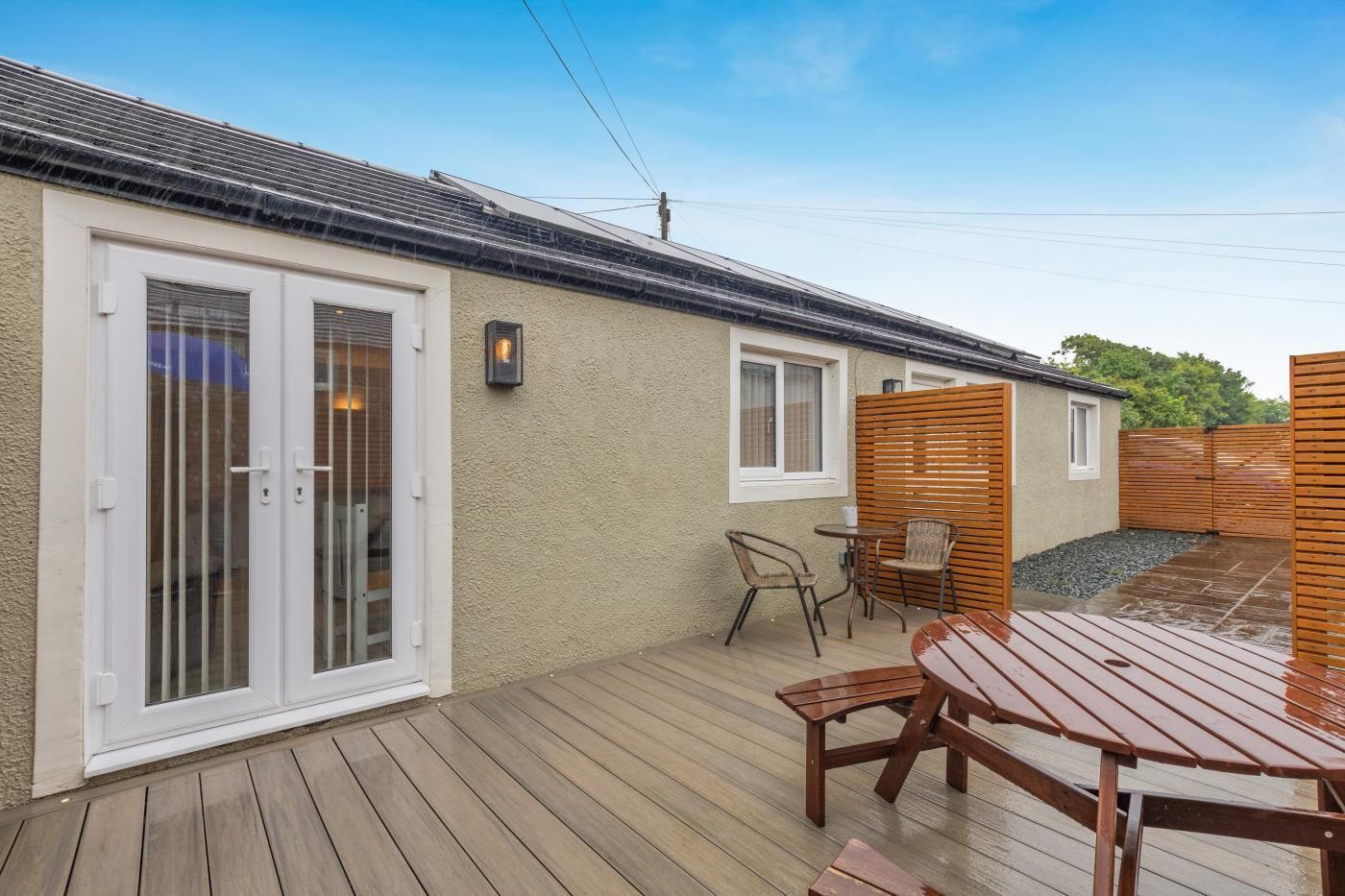 A patio with a wooden deck, a table, chairs, a small garden area, and a building with a sliding glass door and windows.
