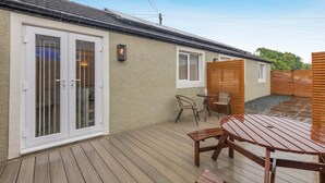 Un patio con terraza de madera, una mesa, sillas, un pequeño jardín y un edificio con puerta corredera de vidrio y ventanas.