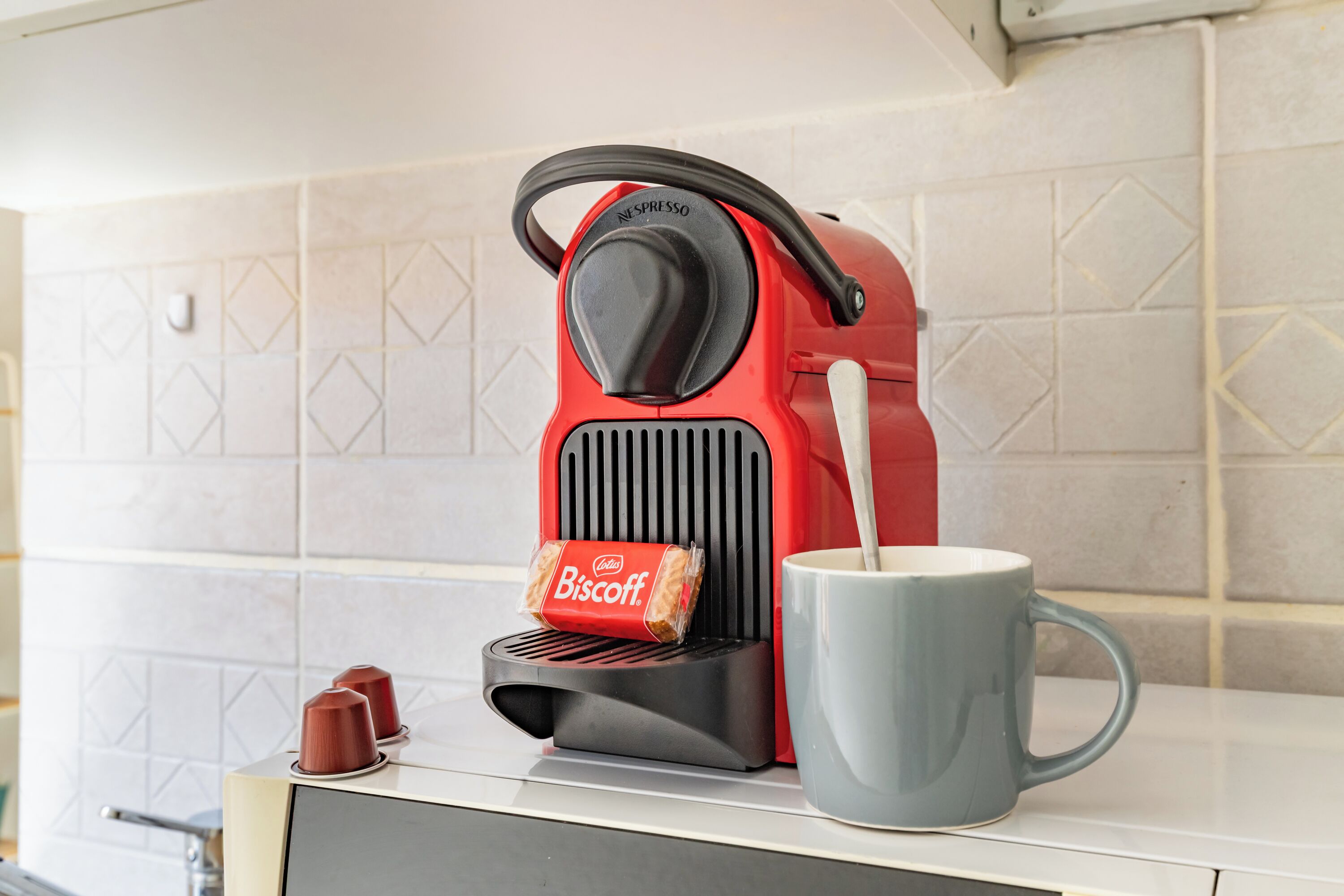 Una máquina de café Nespresso roja con una bolsa de galletas Lotus y una taza azul sobre una encimera de cocina.
