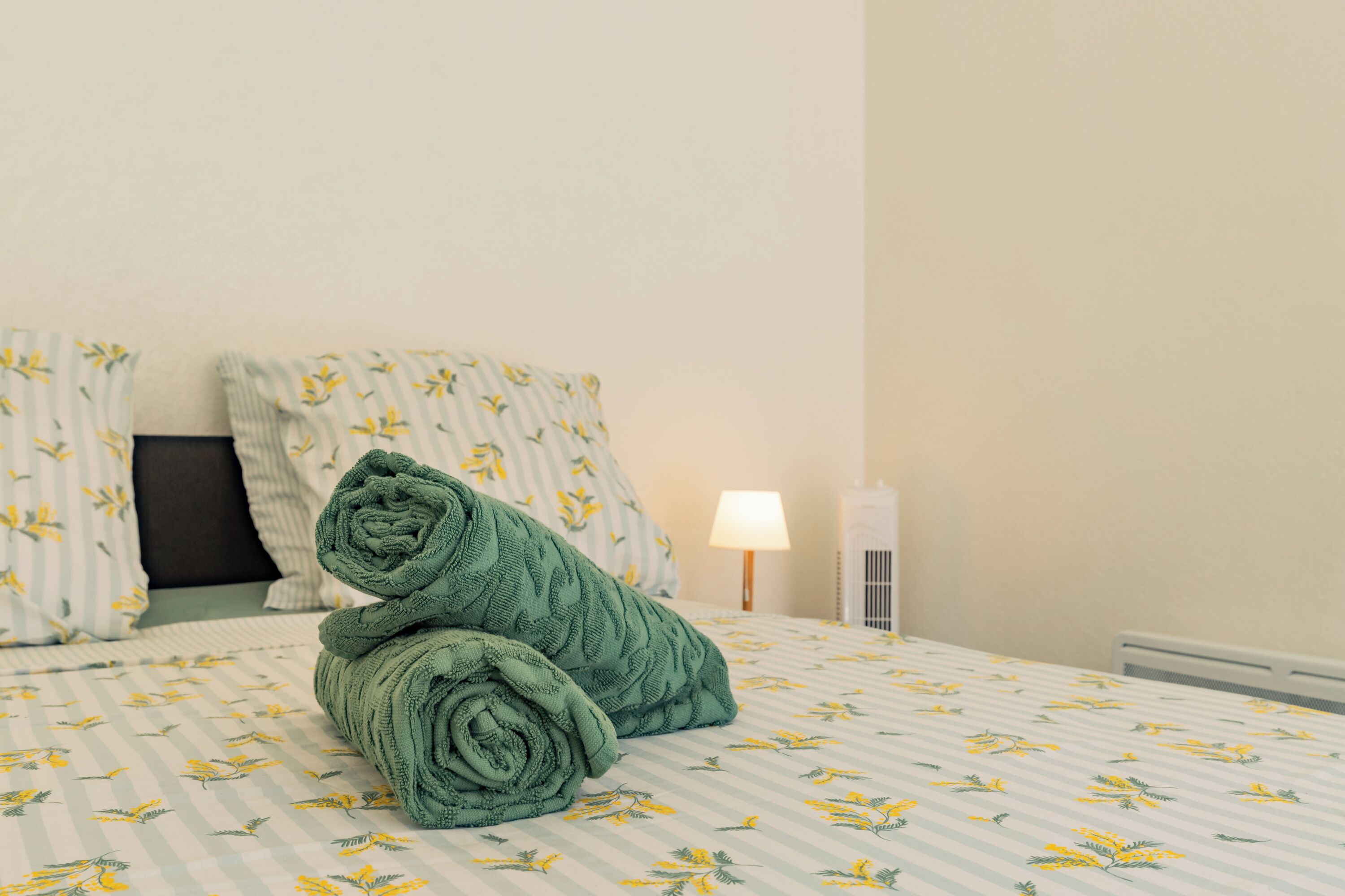 Ein ordentlich bezogenes Bett mit floralem Überwurf und Kissen, ein aufgerolltes grünes Handtuch und eine Nachttischlampe.