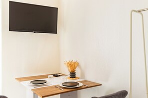 Ein Zimmer mit einem an der Wand befestigten Flachbildfernseher, einem Holztisch mit Geschirr und Besteck sowie einer dekorativen Vase mit Trockenpflanzen.