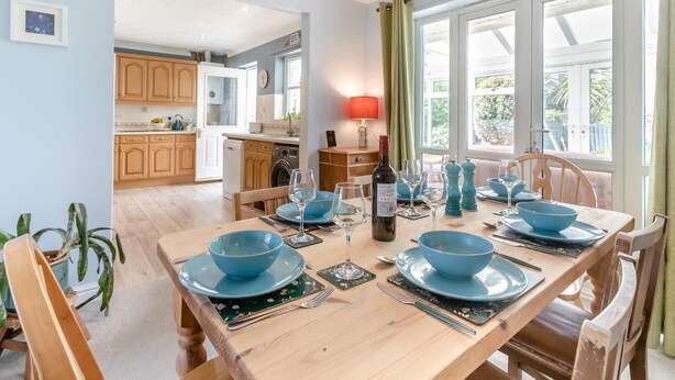 A dining area with a wooden table set for four, a kitchen in the background, and a view of the outdoors. - 3 Bed in Westward Ho (oc-s42079) (Westward Ho!)