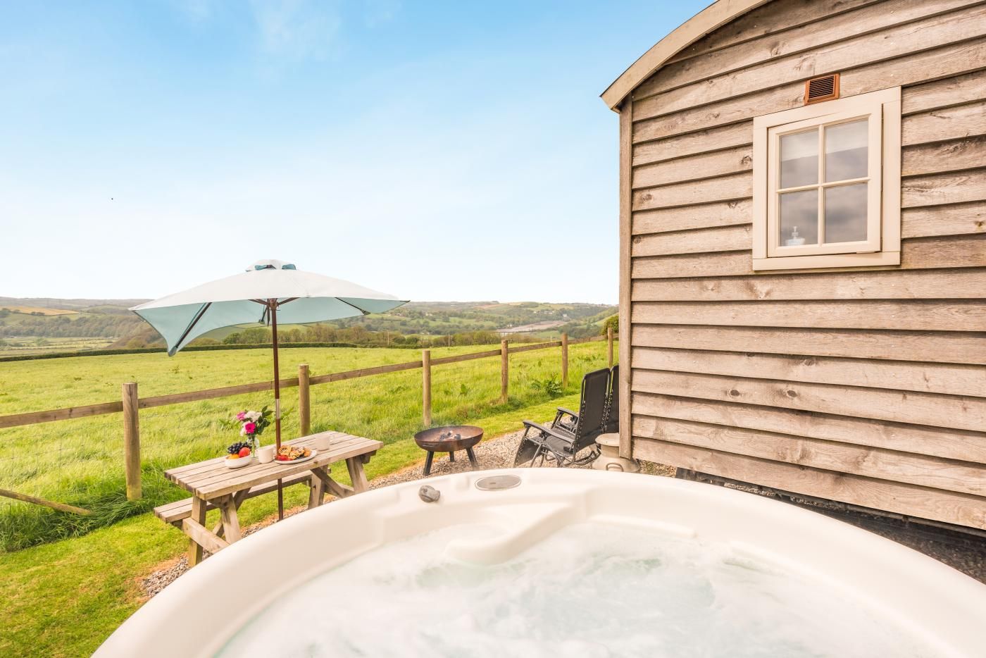 Eine Holzhütte mit Whirlpool, Picknicktisch und Sonnenschirm mit Blick auf eine Wiese.
