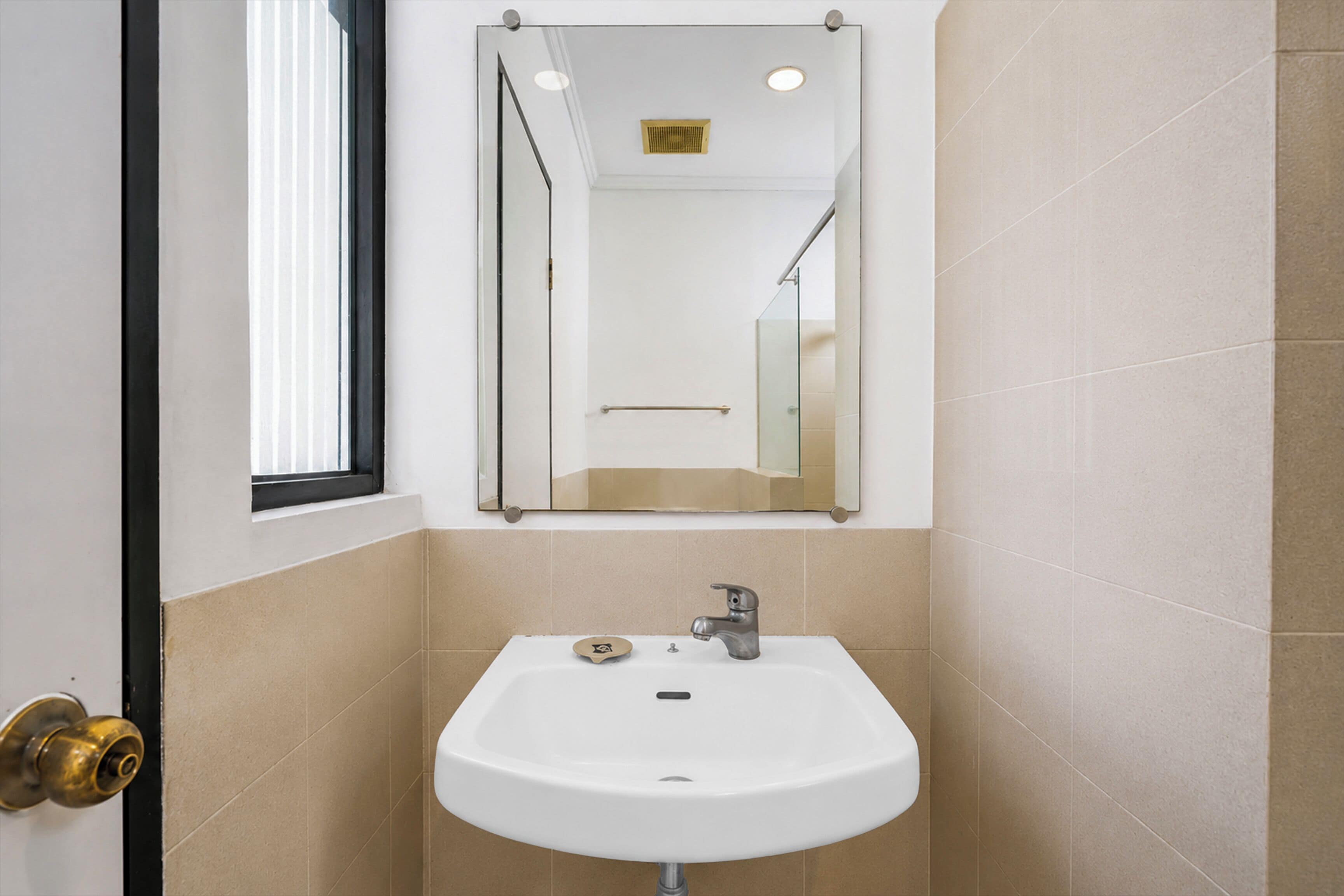 Un baño con lavamanos blanco, un espejo y baldosas color beige.