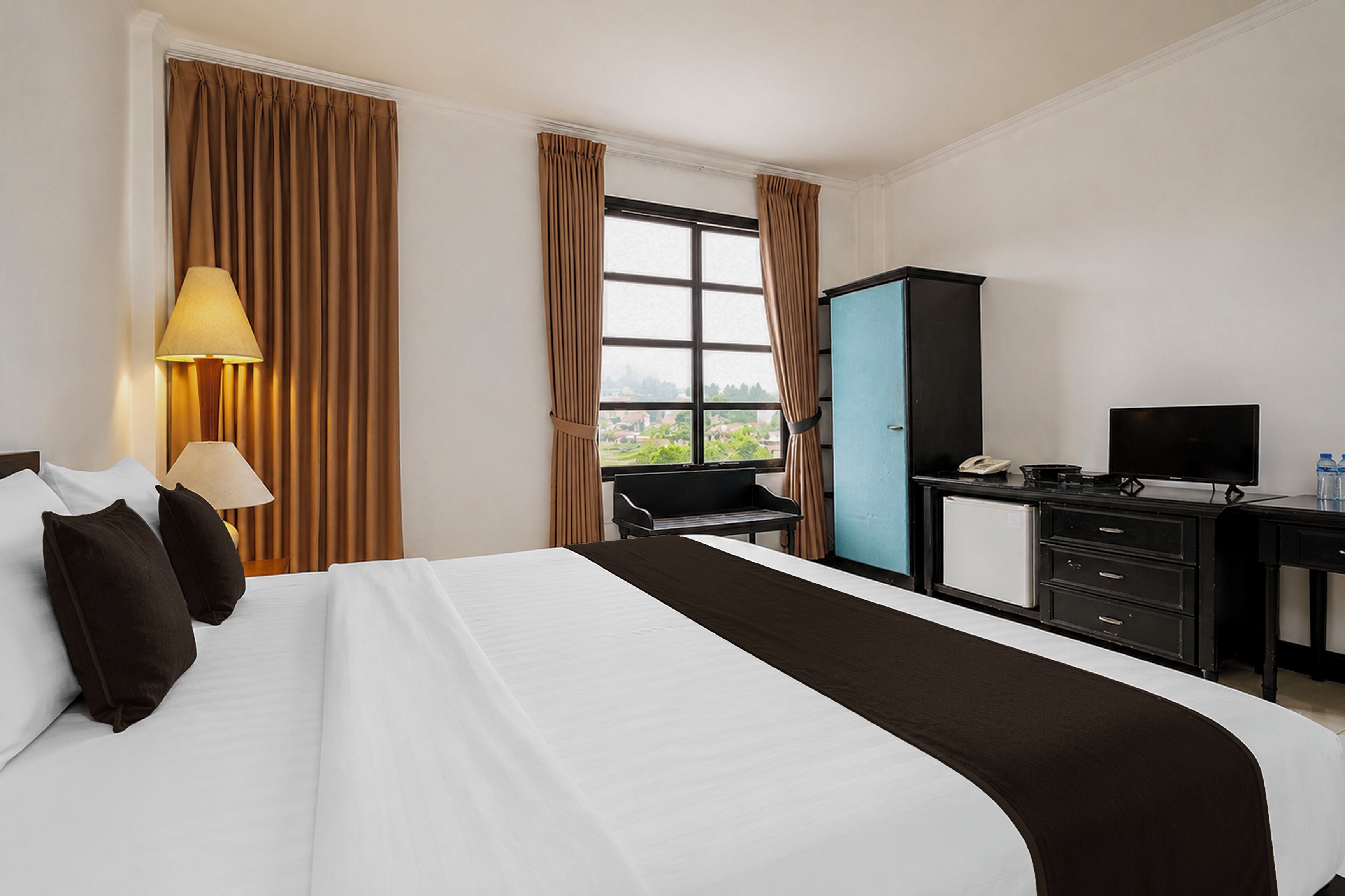 Quarto de hotel com cama, escrivaninha, cadeira, televisão e janela com cortinas.
