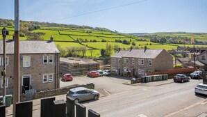 A rural street with stone buildings, parked cars, and a green field in the background.