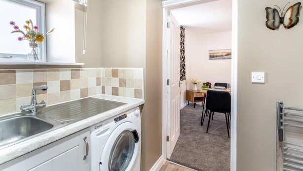 A kitchen with a washing machine, a sink, and a window with flowers. There is a dining area with a table and chairs.