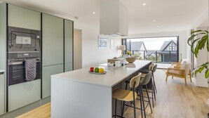 A modern kitchen with a central island, bar stools, and a view of a house through large windows. - 5 Bed in Ogmore-by-Sea (oc-o42632) (Ogmore-by-Sea)