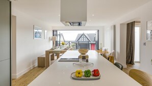 A modern kitchen with a central island, wooden flooring, and a view of a house through large windows. - 5 Bed in Ogmore-by-Sea (oc-o42632) (Ogmore-by-Sea)