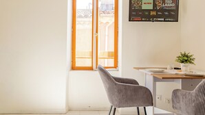 Quarto com janela, duas cadeiras, escrivaninha com computador e televisão na parede exibindo um programa.