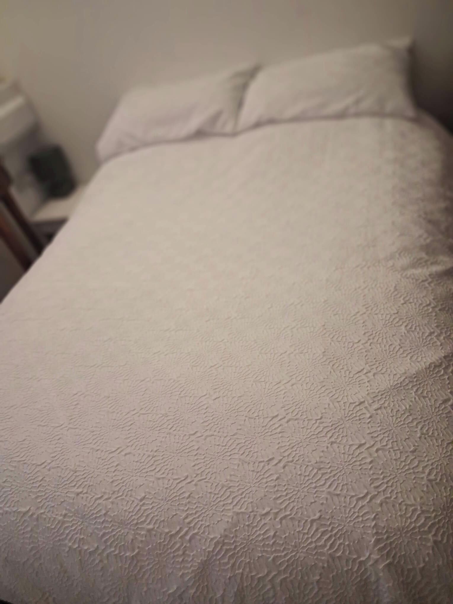 Uma cama com cobertura branca texturizada e um travesseiro.