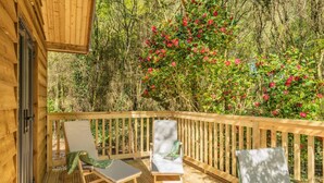 Eine Holzterrasse mit zwei Liegestühlen und einem Tisch, umgeben von dichtem Grün und roten Blüten.