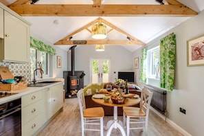 A cozy kitchen with a wood-burning stove, a dining table with chairs, and a TV in the background. - 1 Bed in Buckland Filleigh (oc-h41749) (Buckland Filleigh)