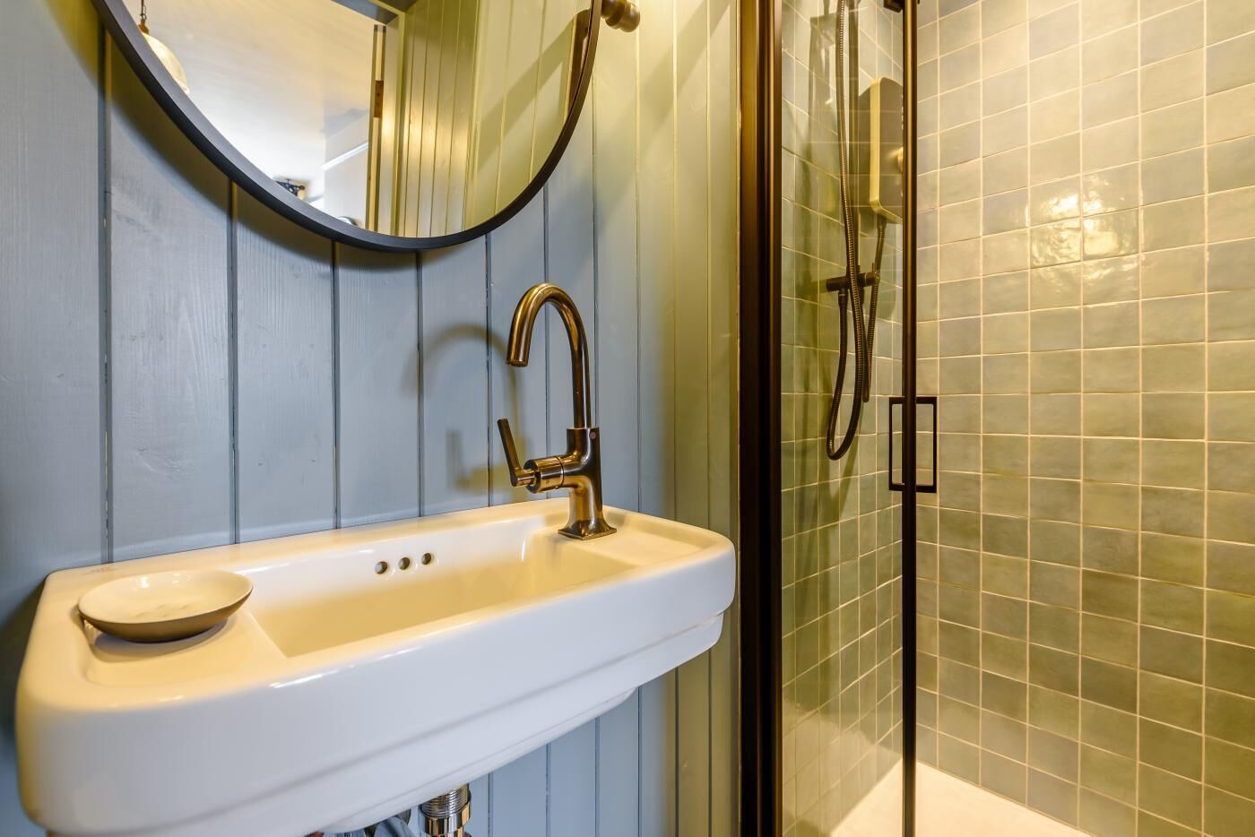 A bathroom with a white sink, a gold faucet, a round mirror, and a glass shower enclosure.