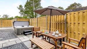 Een terras met een hottub, houten tafel, stoelen en een parasol.