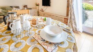 Uma área de refeições com mesa posta para o café da manhã, uma cafeteira, uma TV fixada na parede e vista para o exterior através de uma janela.