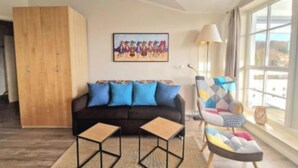 Een woonkamer met een bank, twee bijzettafels, een kleurrijke fauteuil, een houten garderobe en een schilderij aan de muur.