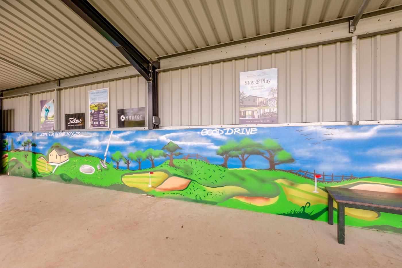 A covered outdoor area with a mural of a golf course, a sign for 'GoodDrive,' and several other signs advertising local businesses.
