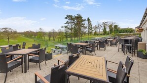 An outdoor patio with multiple tables and chairs, a view of a golf course, and a building with a hanging flower basket. - 1 Bed in Lancashire (oc-w37869) (Bentham)