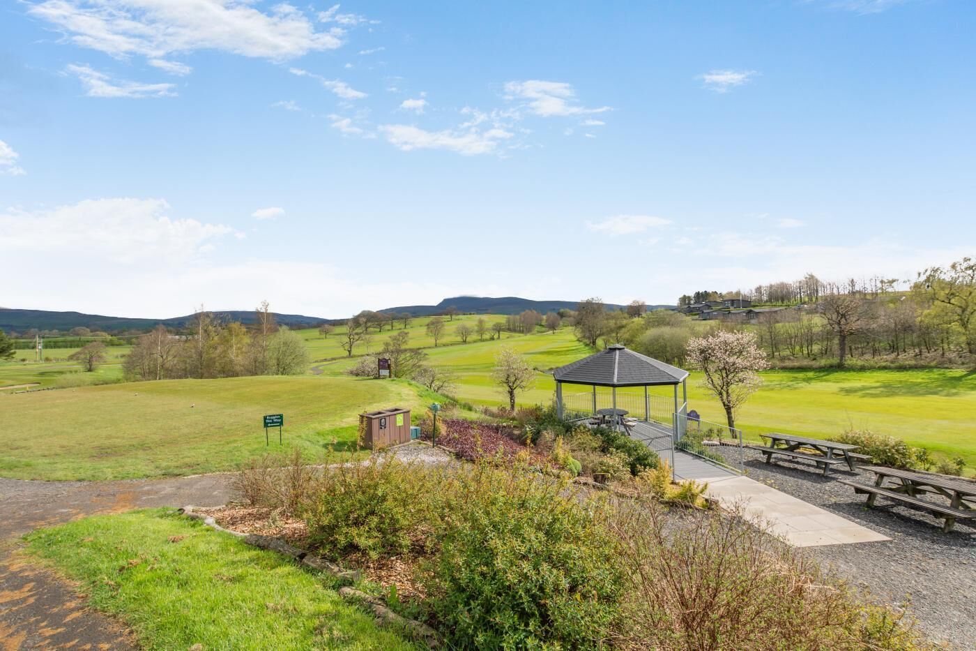 A rural landscape with a gazebo, picnic tables, and a signpost.