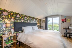 Ein Schlafzimmer mit einem großen Bett, einem Nachttisch, einem Schreibtisch und Blick auf Grün durch ein Fenster.