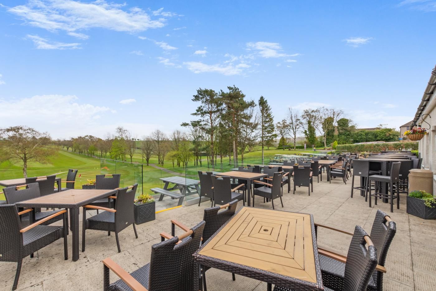 An outdoor patio with multiple tables and chairs, a view of a golf course, and a building with a hanging flower basket.
