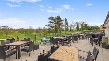 An outdoor patio with multiple tables and chairs, a view of a golf course, and a building with a hanging flower basket.
