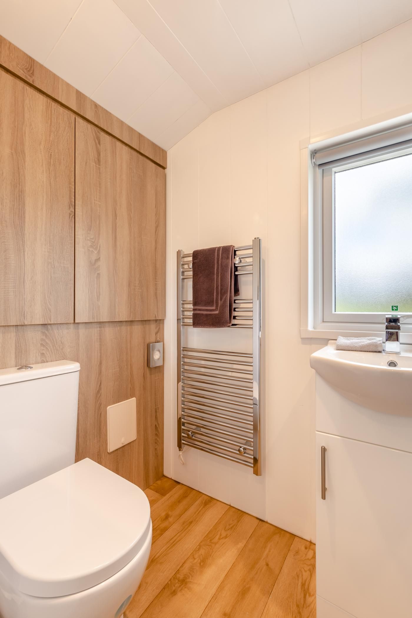 A bathroom with a white toilet, wooden cabinets, a towel rack, and a window.