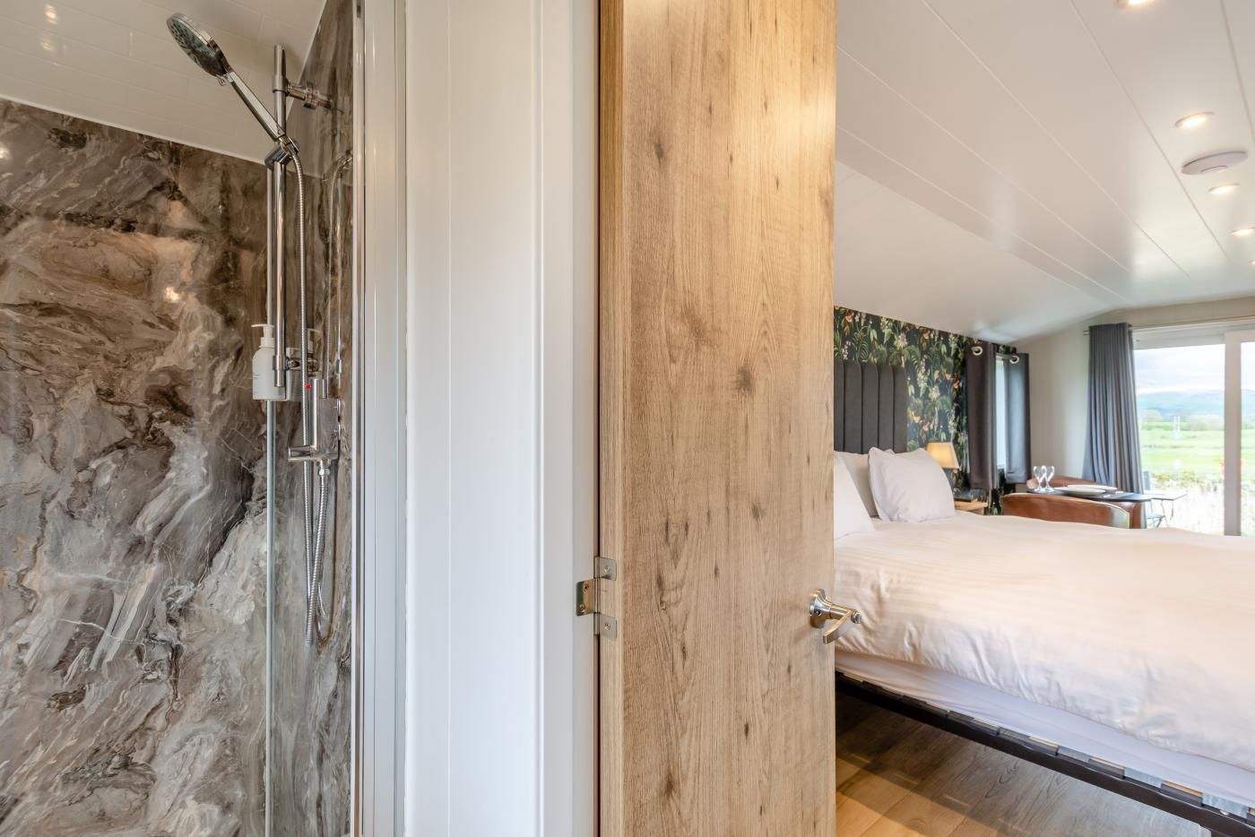 A bedroom with a bed, a wooden door, a shower with a glass enclosure, and a view of the outdoors.