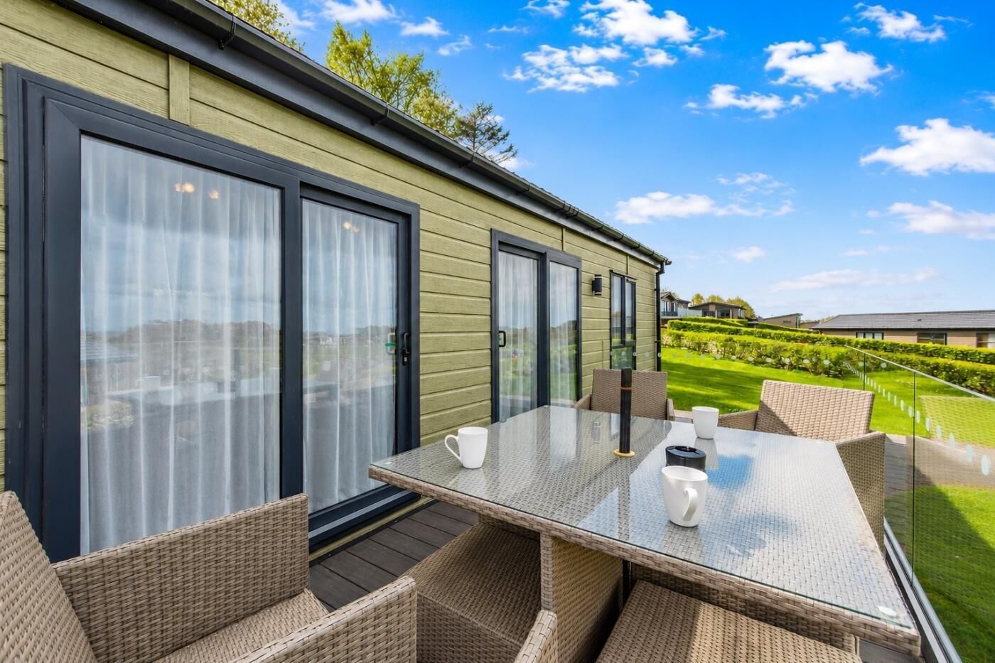 Un espace de restauration extérieur moderne, avec des meubles en osier et une table en verre, offrant une vue sur un jardin bien entretenu et un bâtiment situé au loin.