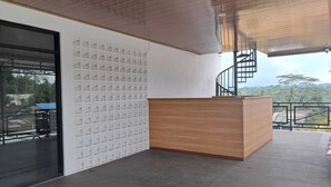 A modern reception desk with a wooden finish, a white wall with a geometric pattern, and a view of a mountainous landscape through a glass door.