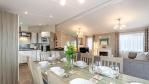 A modern dining area with a wooden table set for four, a kitchen in the background, and a living area with a fireplace. - 4 Bed in Bentham (oc-w37870) (Bentham)