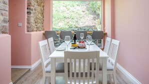 Un comedor con pared de piedra, un ventanal con vista a follaje, sillas blancas y una mesa lista para dos.