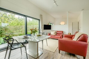 A modern living room with a dining table, red leather sofa, and a large window offering a view of greenery.