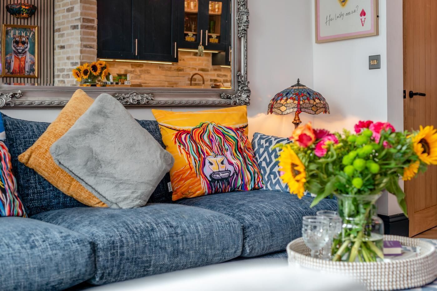 Una sala de estar con un sofá, cojines decorativos, un florero con flores y un espejo.