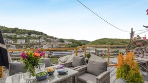 Een terras met rieten meubels, een glazen tafel en uitzicht op een rivier en gebouwen.
