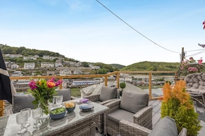 Eine Terrasse mit Korbmöbeln, einem Glastisch und Blick auf einen Fluss sowie Gebäude.
