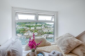 Ein Zimmer mit großem Fenster und Blick auf eine Küstenstadt, ein Bett mit Strickdecke und eine Vase mit Blumen.