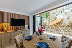 A dining area with a table set for two, a TV mounted on the wall, and a view of a rocky outdoor area with a surfboard. - 4 Bed in Looe (oc-a36906) (Looe)