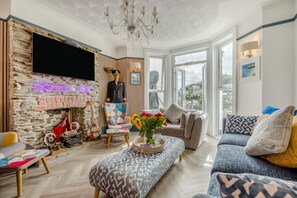 A living room with a stone wall, a flat-screen TV, a chandelier, and a large window offering a city view. - 4 Bed in Looe (oc-a36906) (Looe)